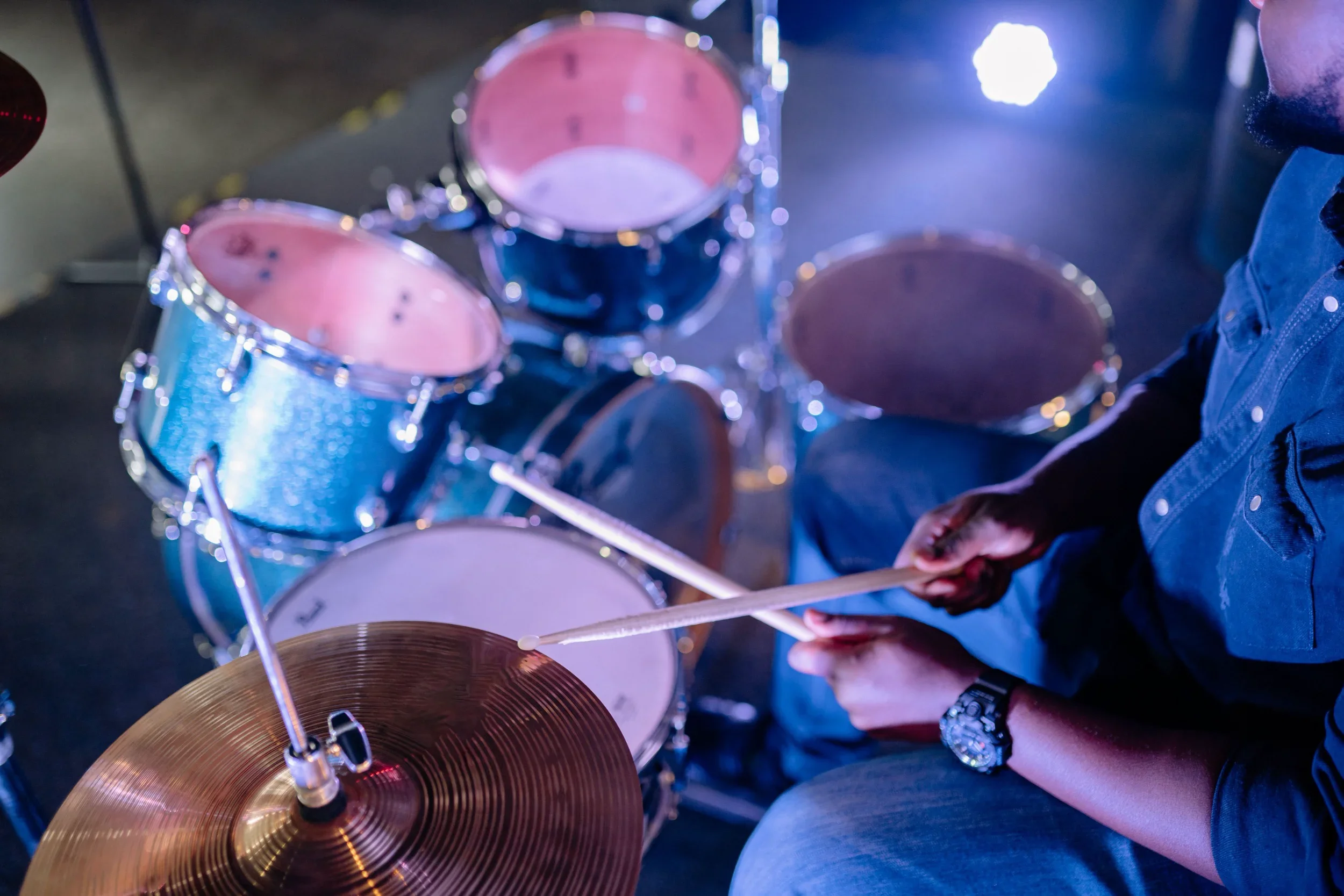 A person playing a blue drum kit with a pair of drumsticks in a dark room illuminated by purple and blue lights.
