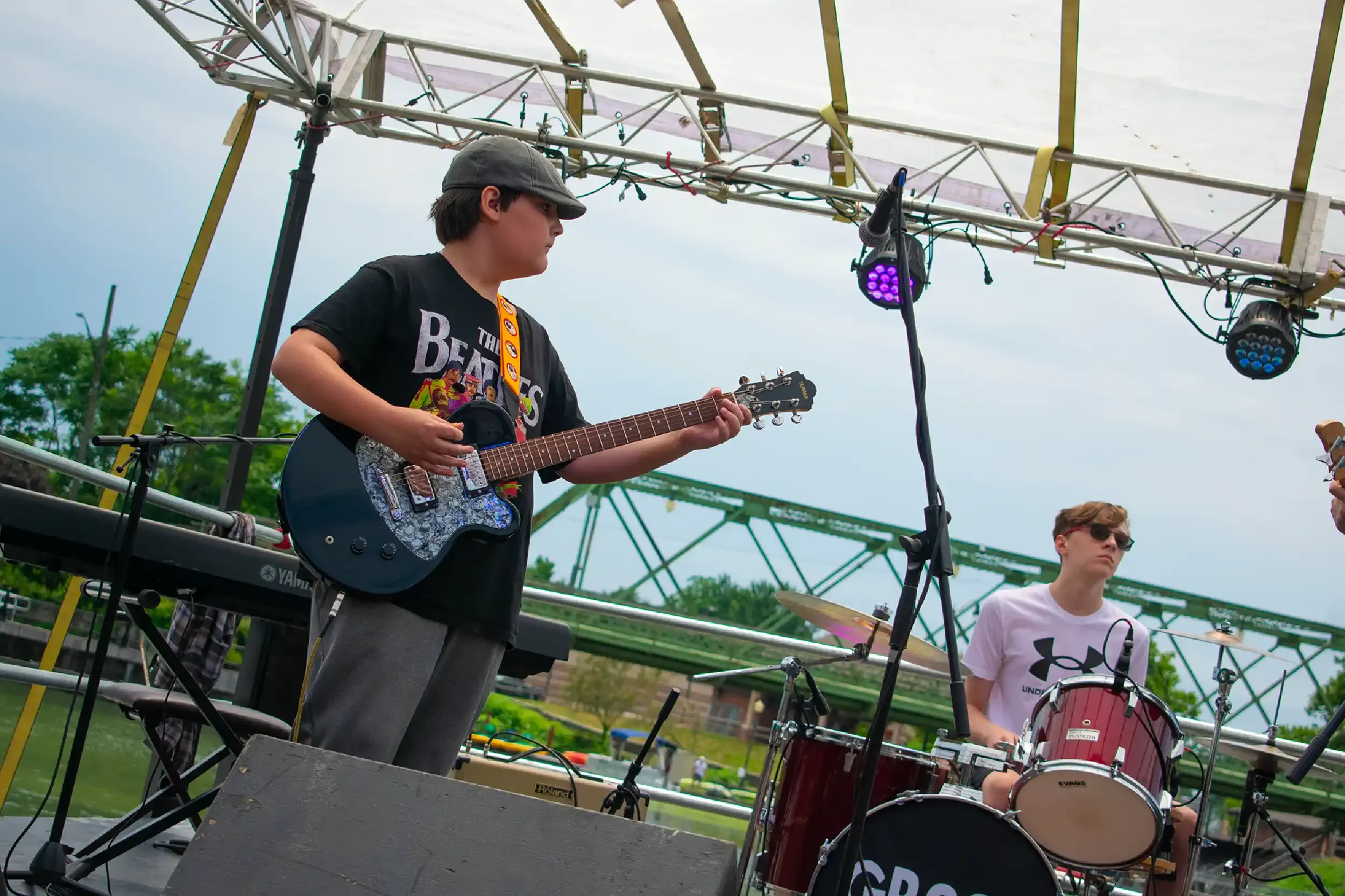 Two young male musicians performing outdoors under a metal stage structure. One is playing an electric guitar and the other is on drums. The sky is partly cloudy and there are trees and a water feature in the background.