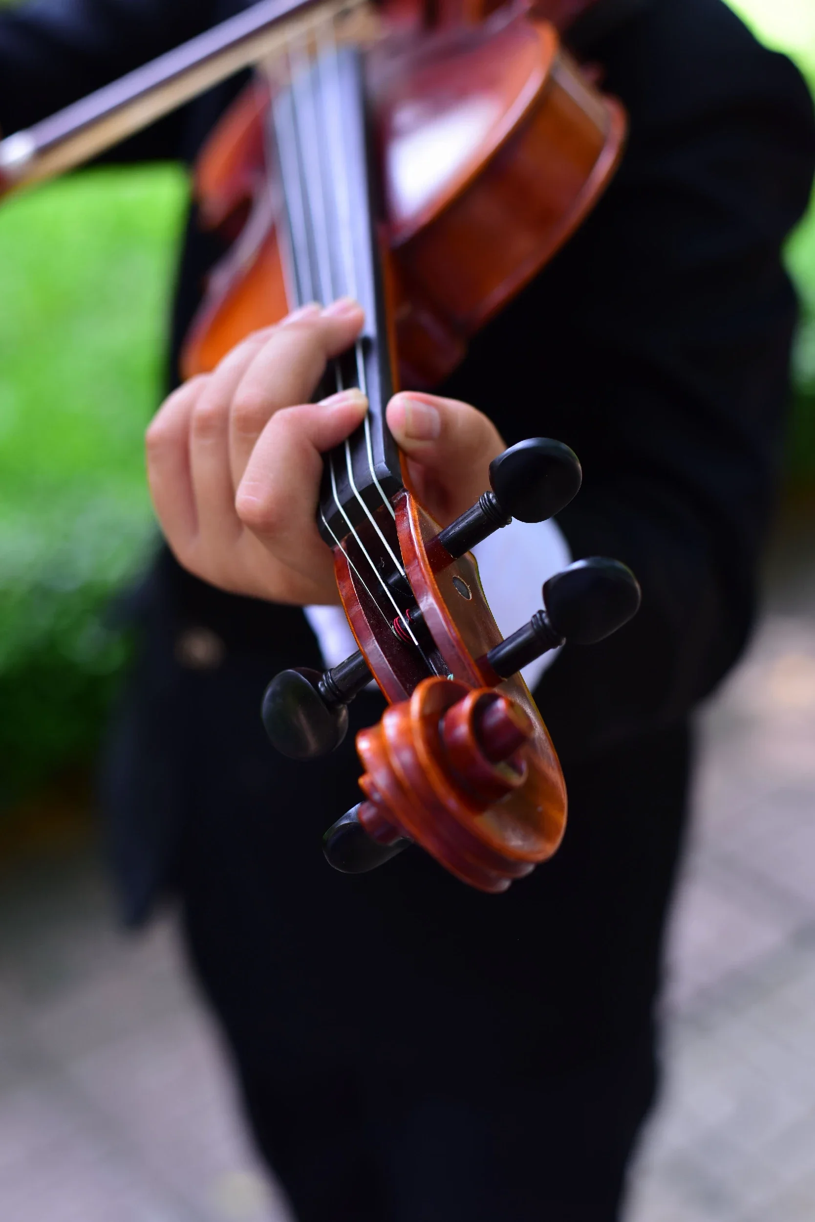 Close-up of a person playing a violin outdoors, focusing on their hand on the strings and the scroll of the violin.