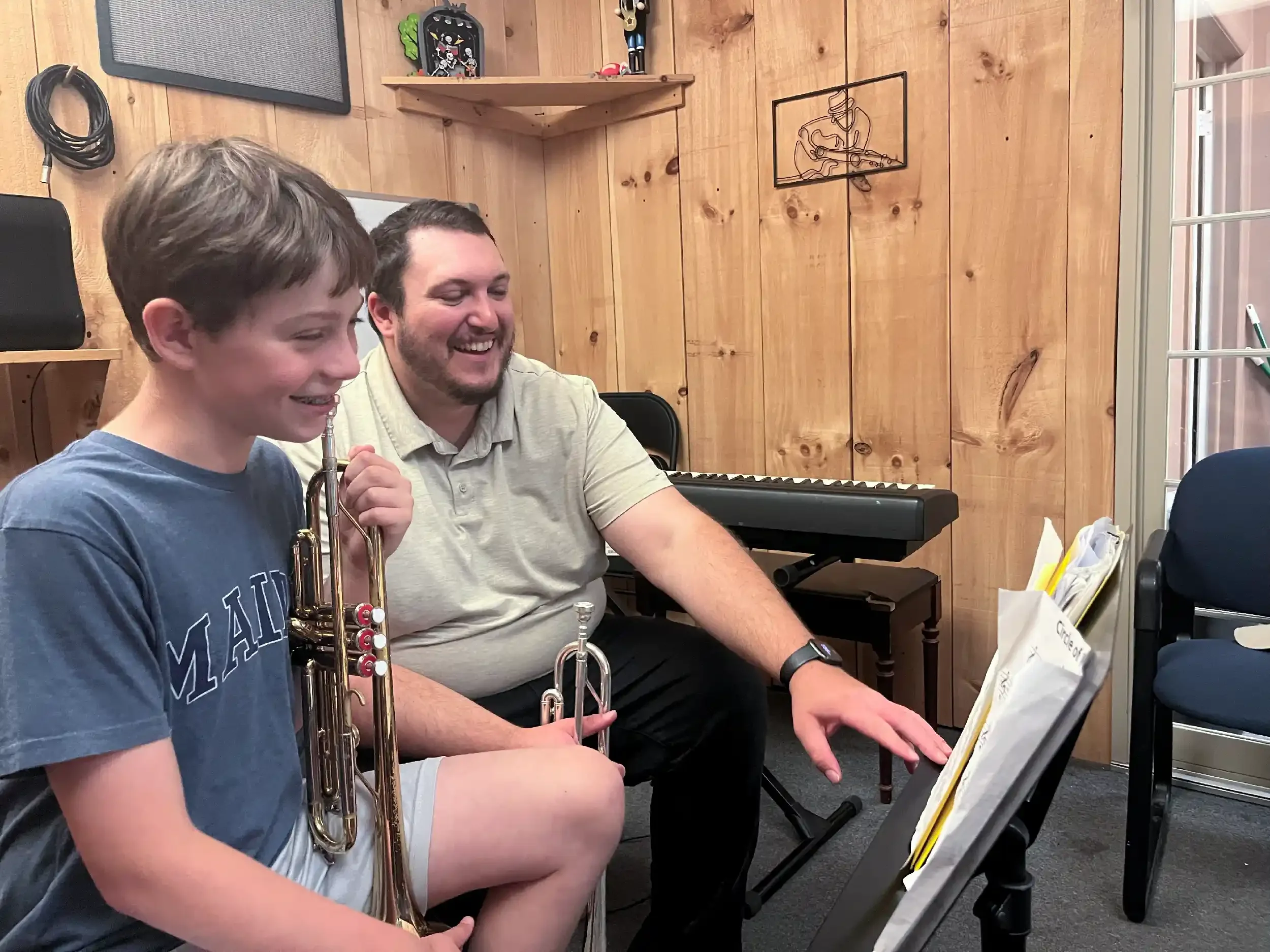 Two males sitting in a music room, one holding a trumpet, both looking at sheet music on a stand, smiling and enjoying playing together.