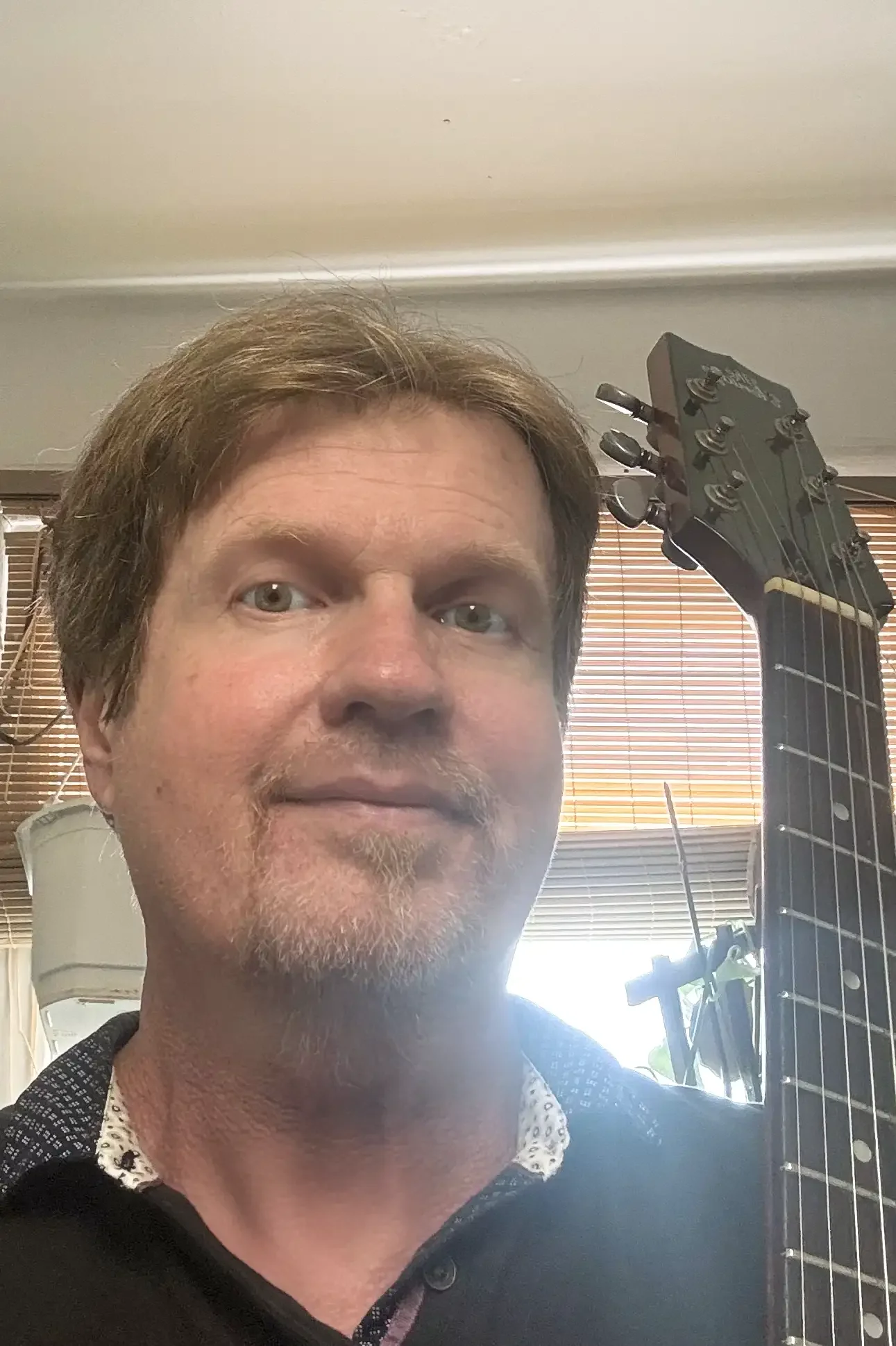 A man with light brown hair, green eyes, and a beard is taking a selfie in a room with blinds, a guitar, and a lamp in the background.