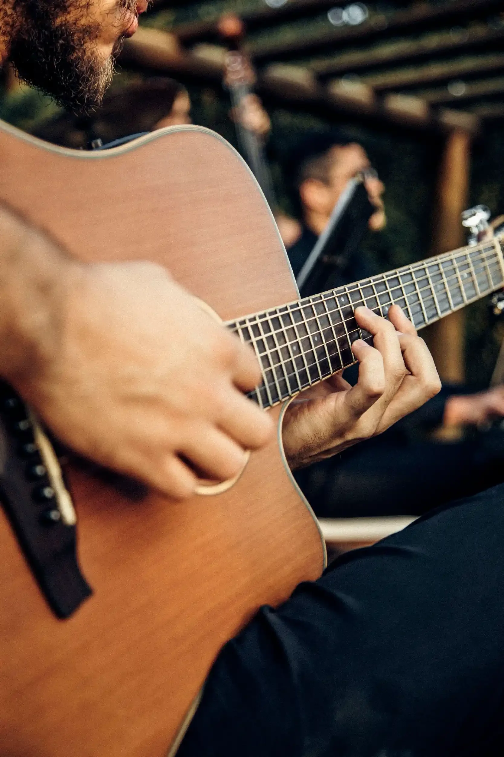 Close-up of a person playing an acoustic guitar with focus on their hand on the fretboard, with two other people in the background also holding guitars, in an outdoor setting.