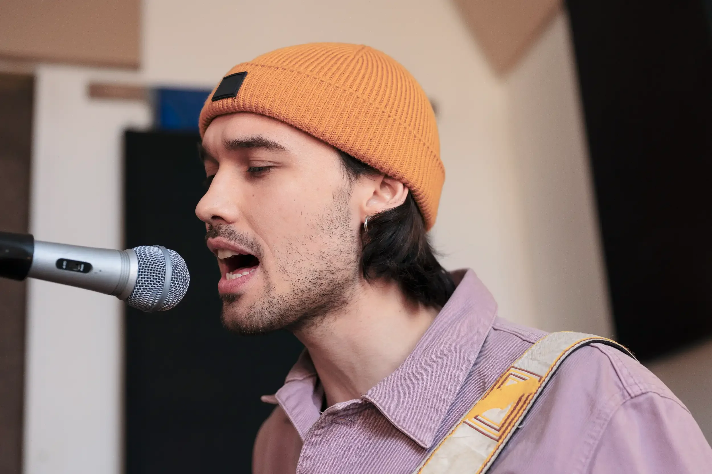 A young man with dark hair, wearing a yellow beanie and a pink shirt, singing into a microphone while playing a guitar, with a blurred background.