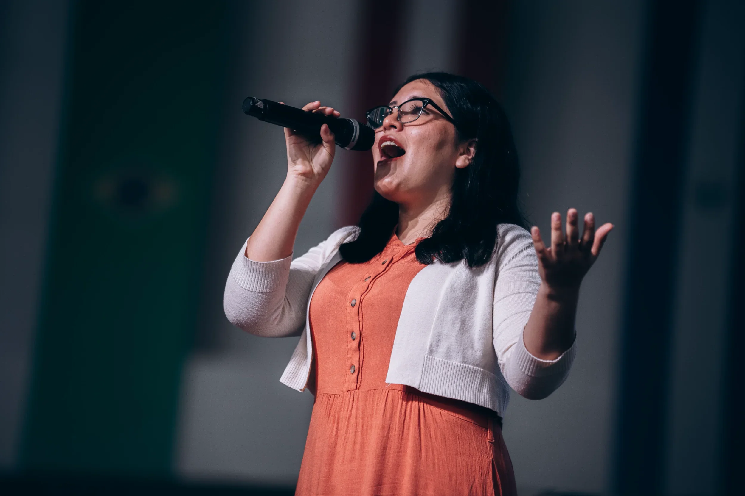 Woman with dark hair and glasses singing passionately into a microphone on stage, wearing an orange dress and a light-colored cardigan.