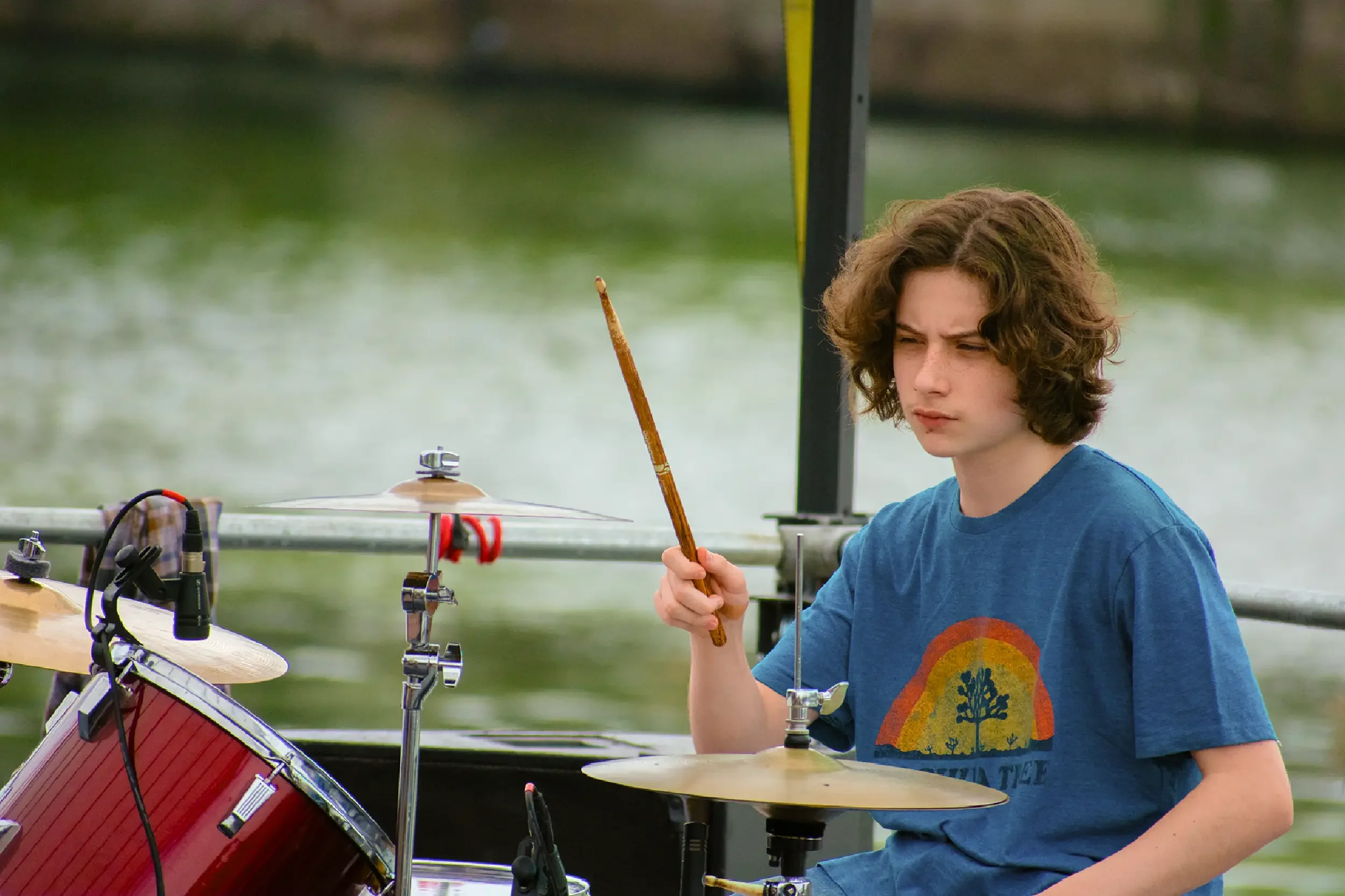 A young boy with curly brown hair playing drums outdoors by a body of water, holding a drumstick with a focused expression.