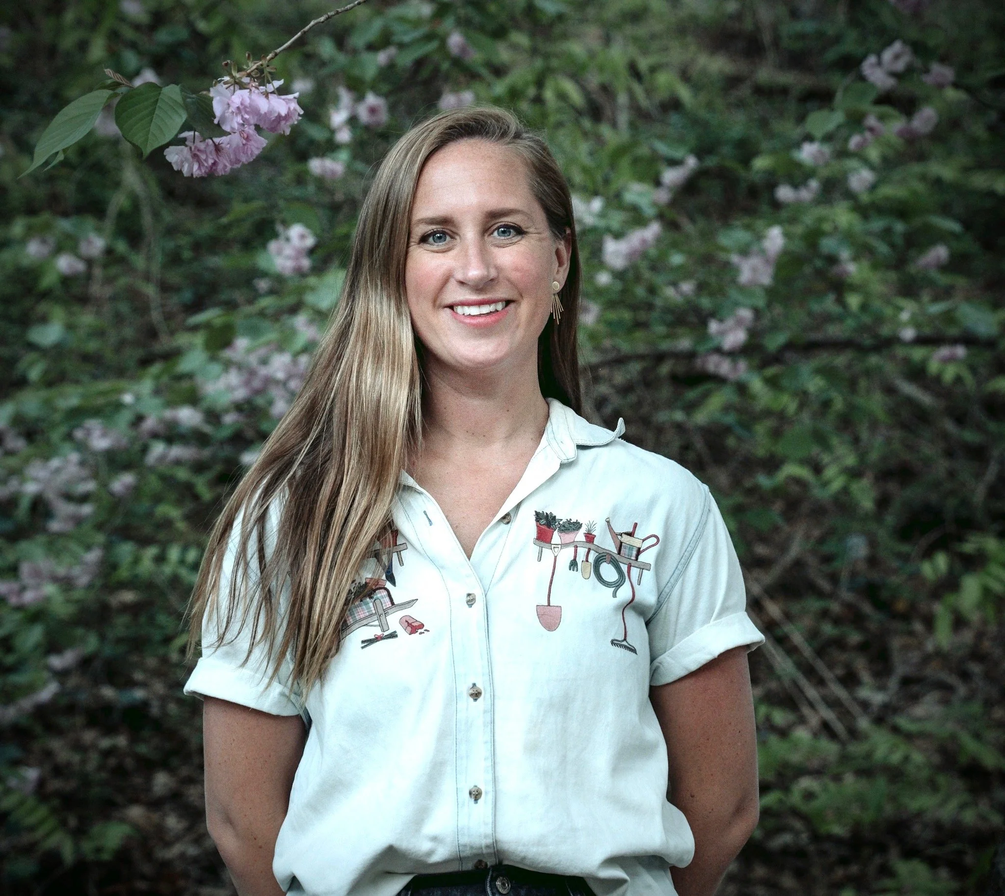 A woman with long blond hair and blue eyes smiling outdoors in front of green foliage and pink flowering branches. She is wearing a white short-sleeve button-up shirt with gardening tools embroidered on it.