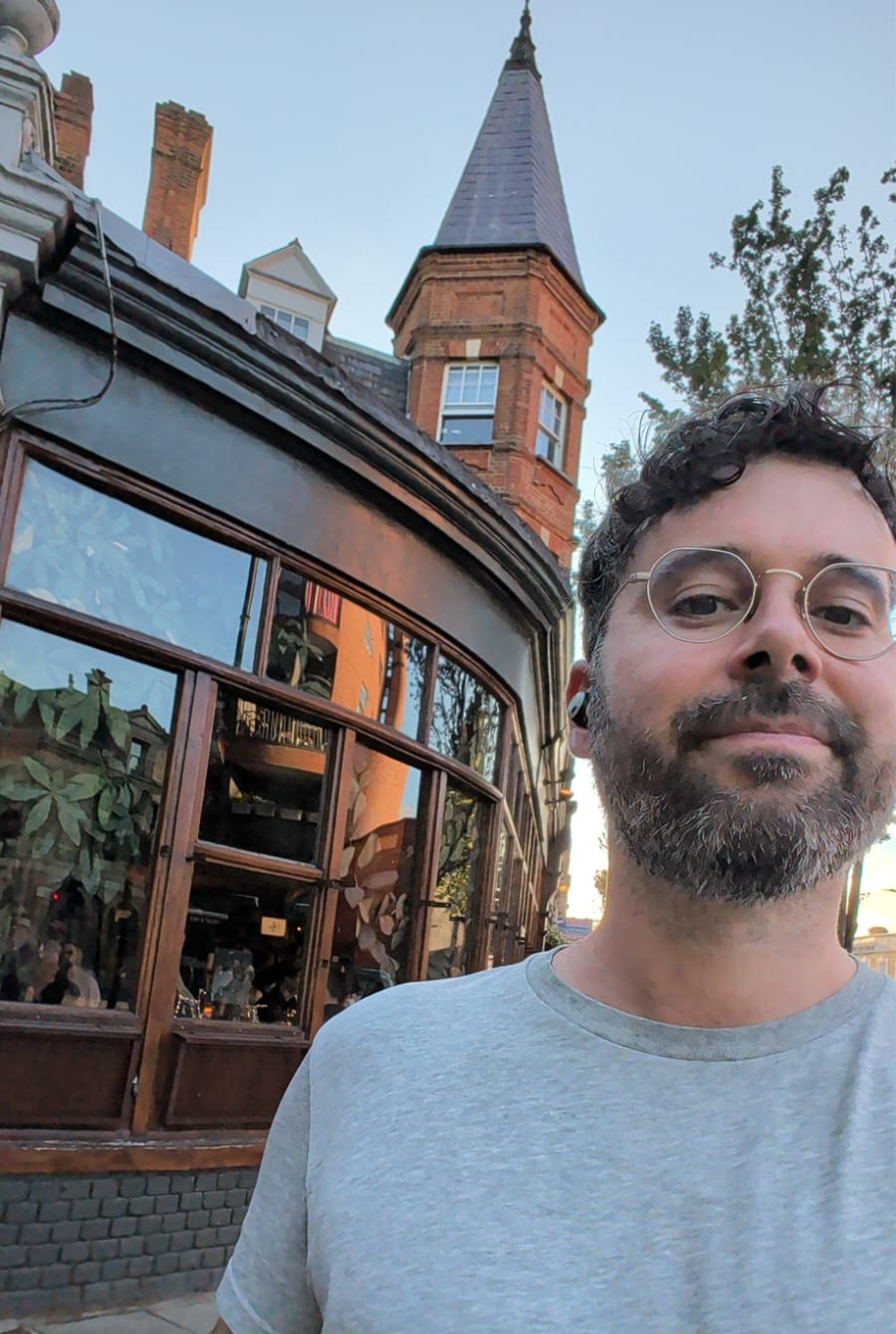 A close-up of a man with black curly hair, glasses, and a beard, wearing a light gray t-shirt, standing outdoors in front of a historic brick building with a tower, during daylight.