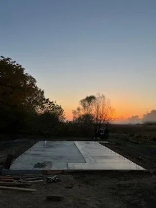 Sunset over open land with trees, showing a concrete or similar surface on the ground, possibly a foundation or slab, with construction materials nearby.