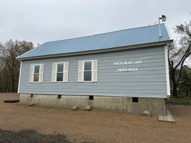 The image shows a tilted little town hall building labeled White Bear Lake Town Hall, with four windows, light blue siding, and a metal roof, on a dirt ground with trees in the background.
