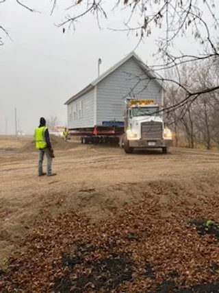 A large white house being transported on a flatbed truck, with one person standing nearby on a muddy ground, and leafless trees in the background on a cloudy day.