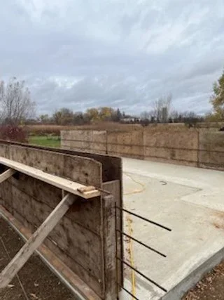 A small, partially built concrete structure with wooden and metal support beams and a wooden fence in the foreground, under a cloudy sky.