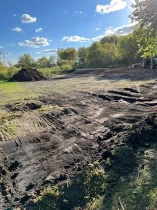 A cleared dirt field with tire tracks, surrounded by trees under a partly cloudy sky.