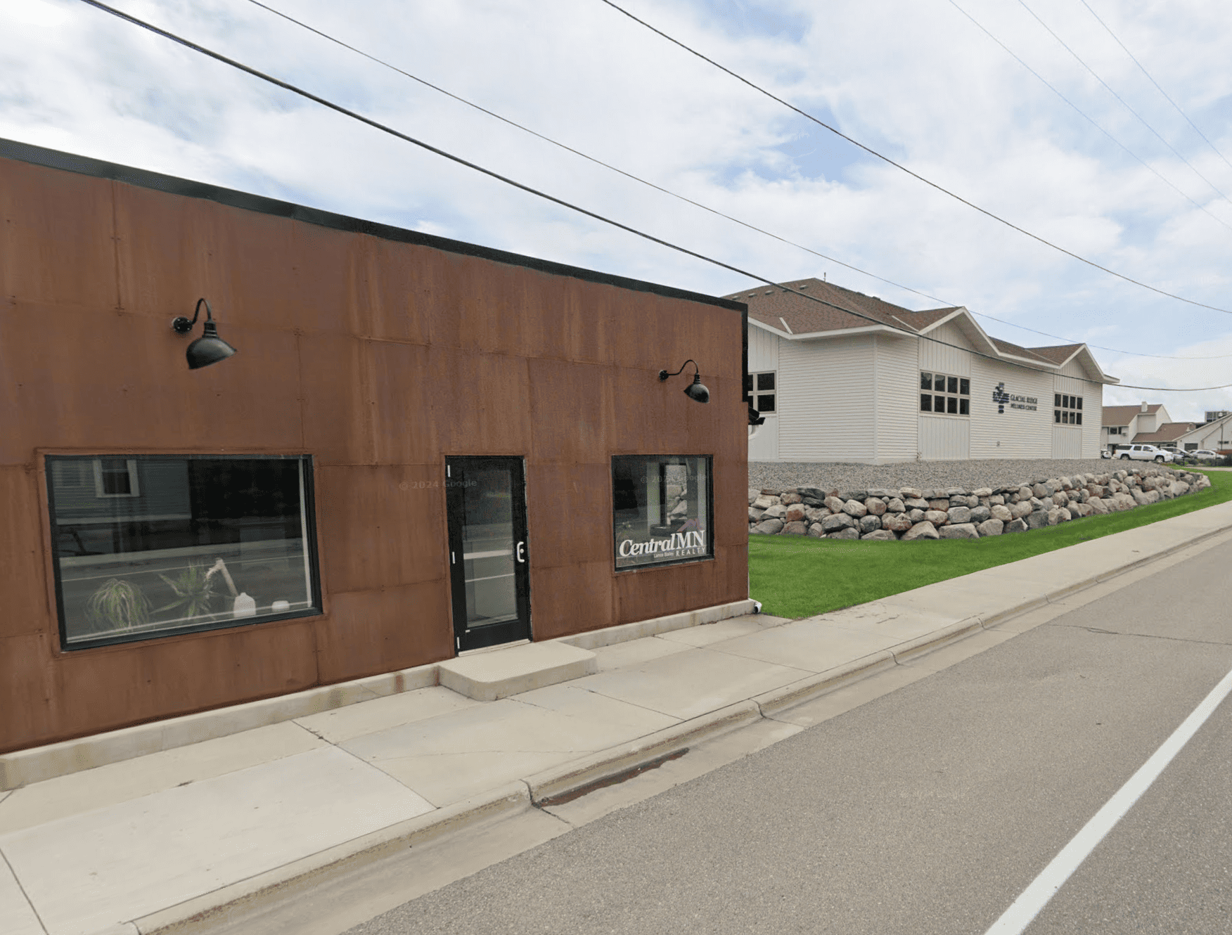 A small commercial building with a brown exterior, two windows, and a door, and a sign that reads 'Central MN Realty.' Next to it is a larger white building with multiple windows and a sign for a church. The sidewalk runs along the street, and there is a landscaped area with large rocks and grass in the background. Power lines are visible in the sky.