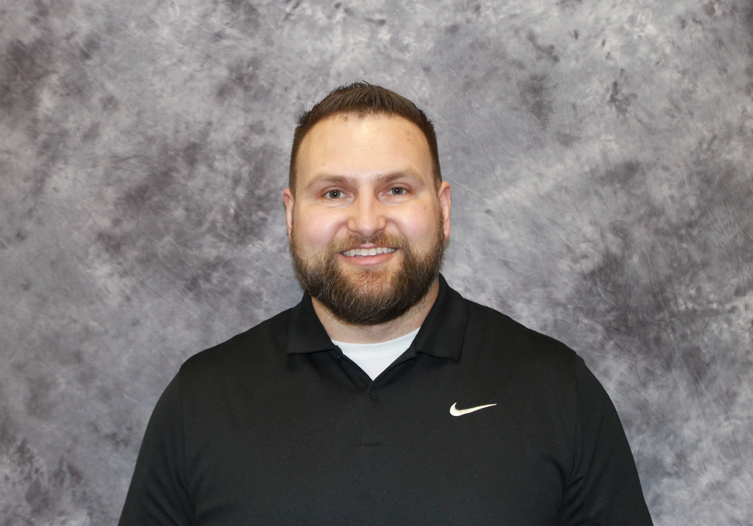 Portrait of a smiling man with a beard wearing a black Nike shirt, standing against a gray textured backdrop.