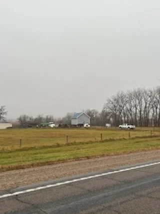 A rural scene with a grassy field, trees in the background, and a few vehicles along a gravel road under an overcast sky.