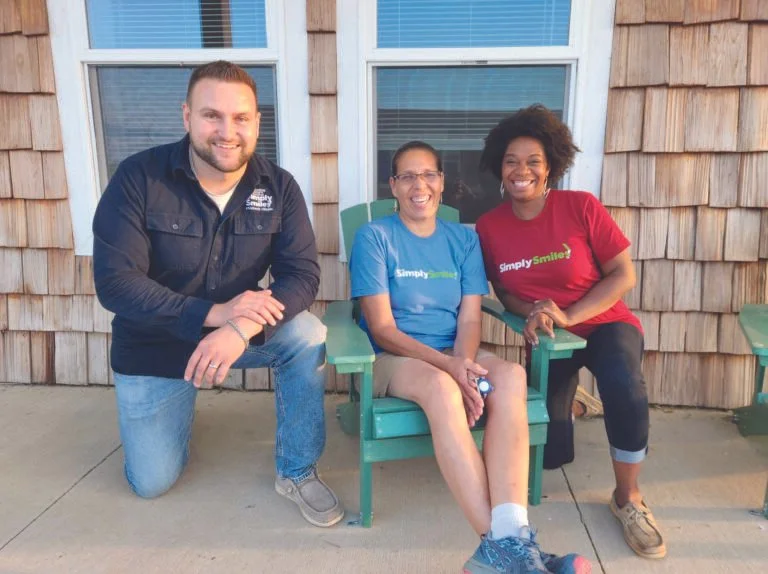 Three people sitting and kneeling on a concrete porch in front of a building with wooden shingles. They are smiling and wearing colorful t-shirts with the logo 'Simply Smiled.'
