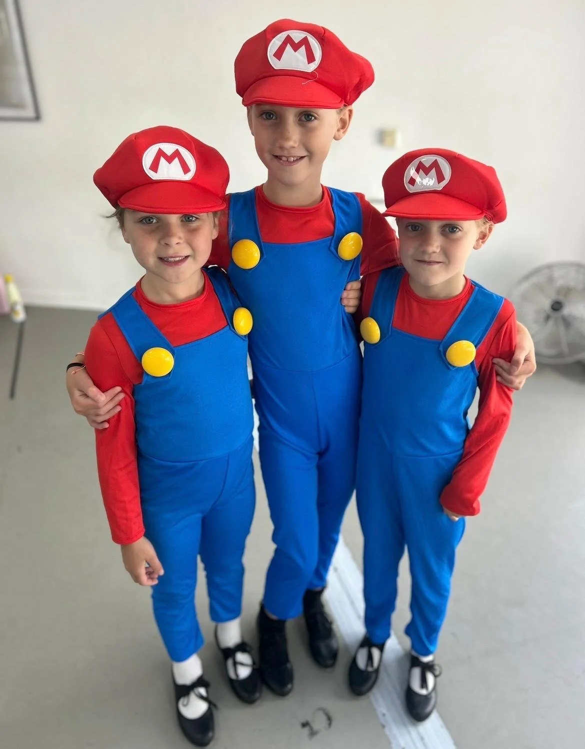 Three children dressed as Mario from Super Mario, wearing red hats with an 'M', red shirts, blue overalls with yellow buttons, and black shoes, standing together with arms around each other.