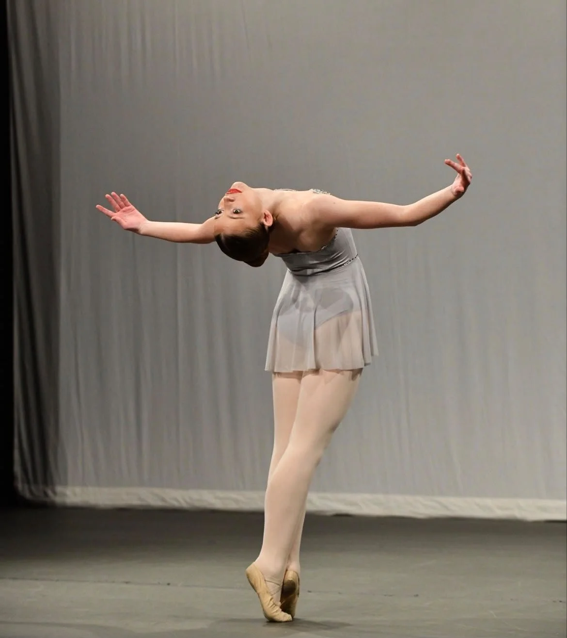A ballerina performing a dance move on stage, wearing a light gray dress and beige tights, with her head tilted back and arms extended.