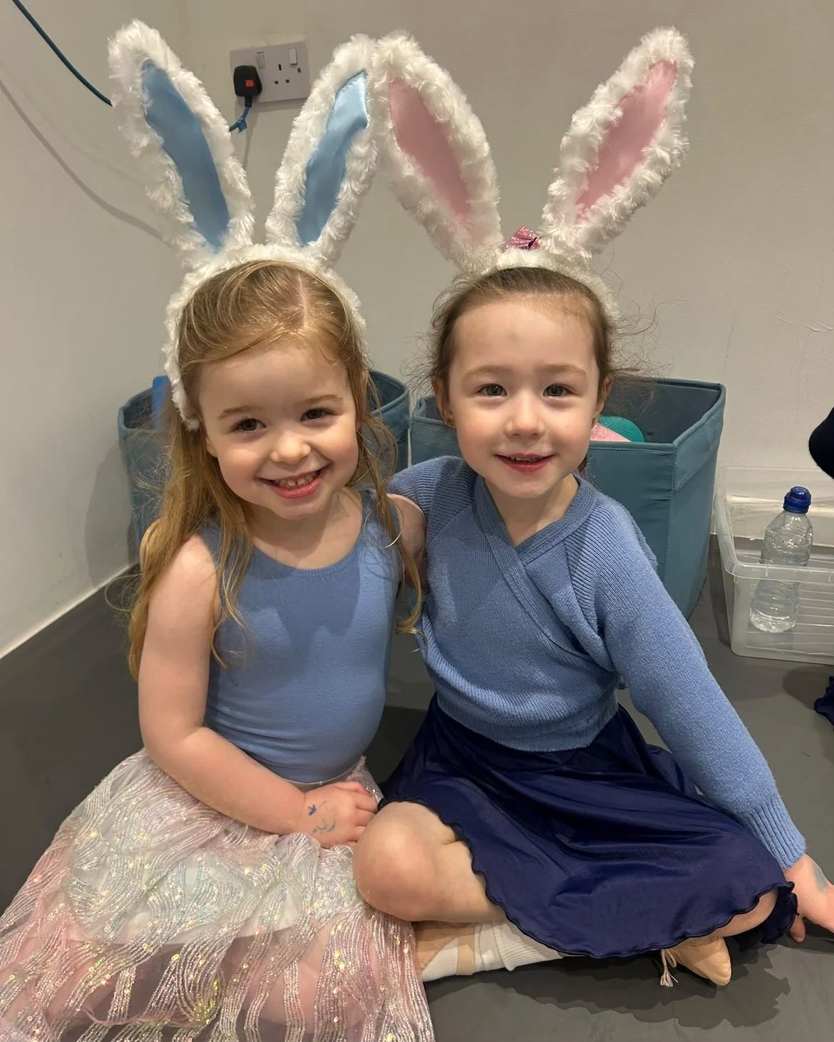 Two young girls wearing bunny ear headbands and light blue clothing, sitting together and smiling indoors.