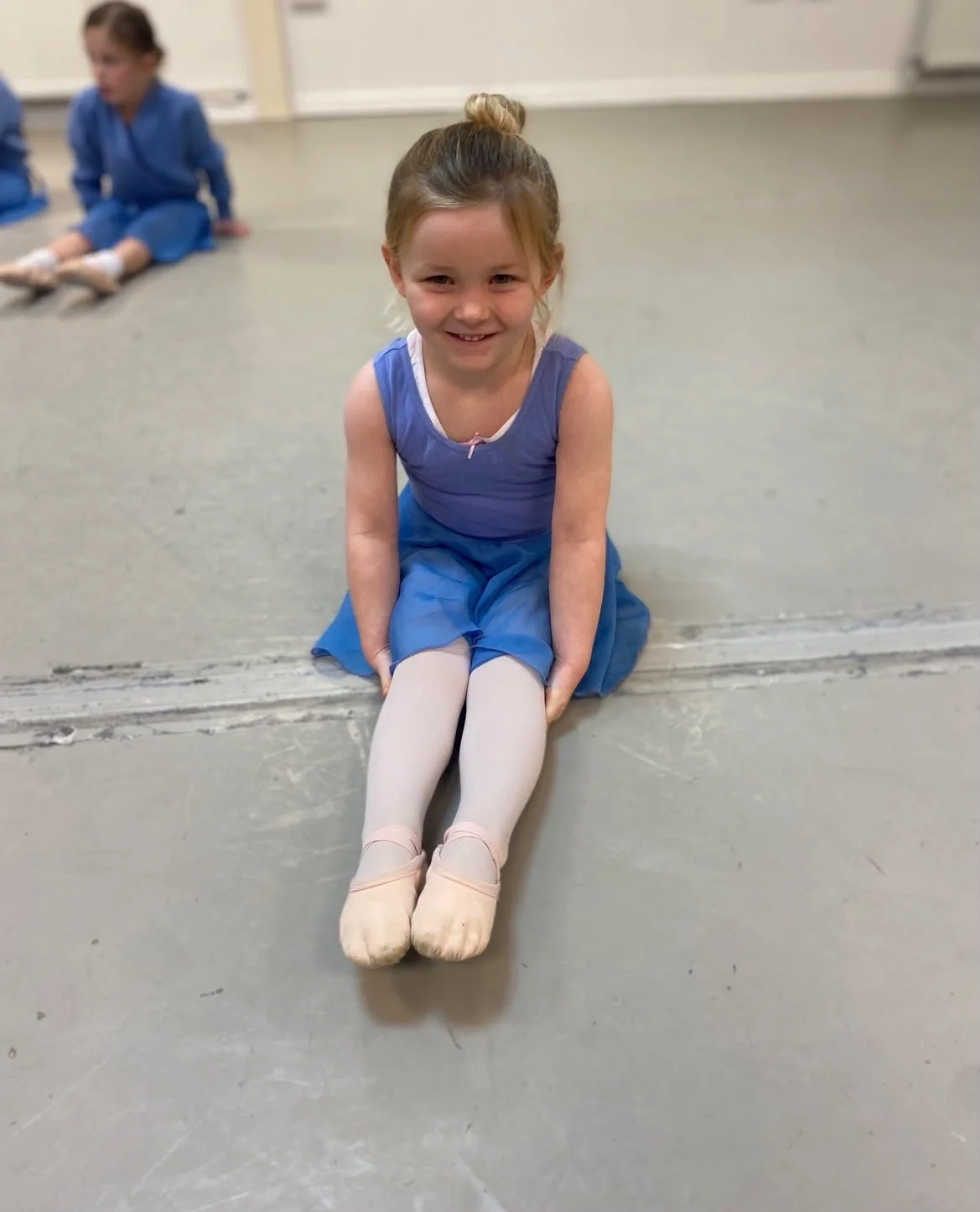 Young girl in a blue dress and ballet slippers sitting on the floor with her legs extended, smiling at the camera, with another girl in a blue outfit in the background.