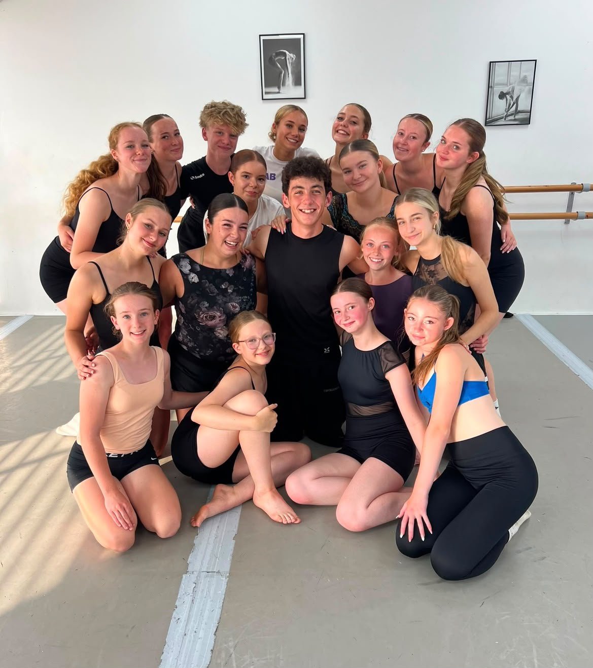 Group of young dancers in a dance studio, posing for a photo with an instructor or teammate, all smiling.