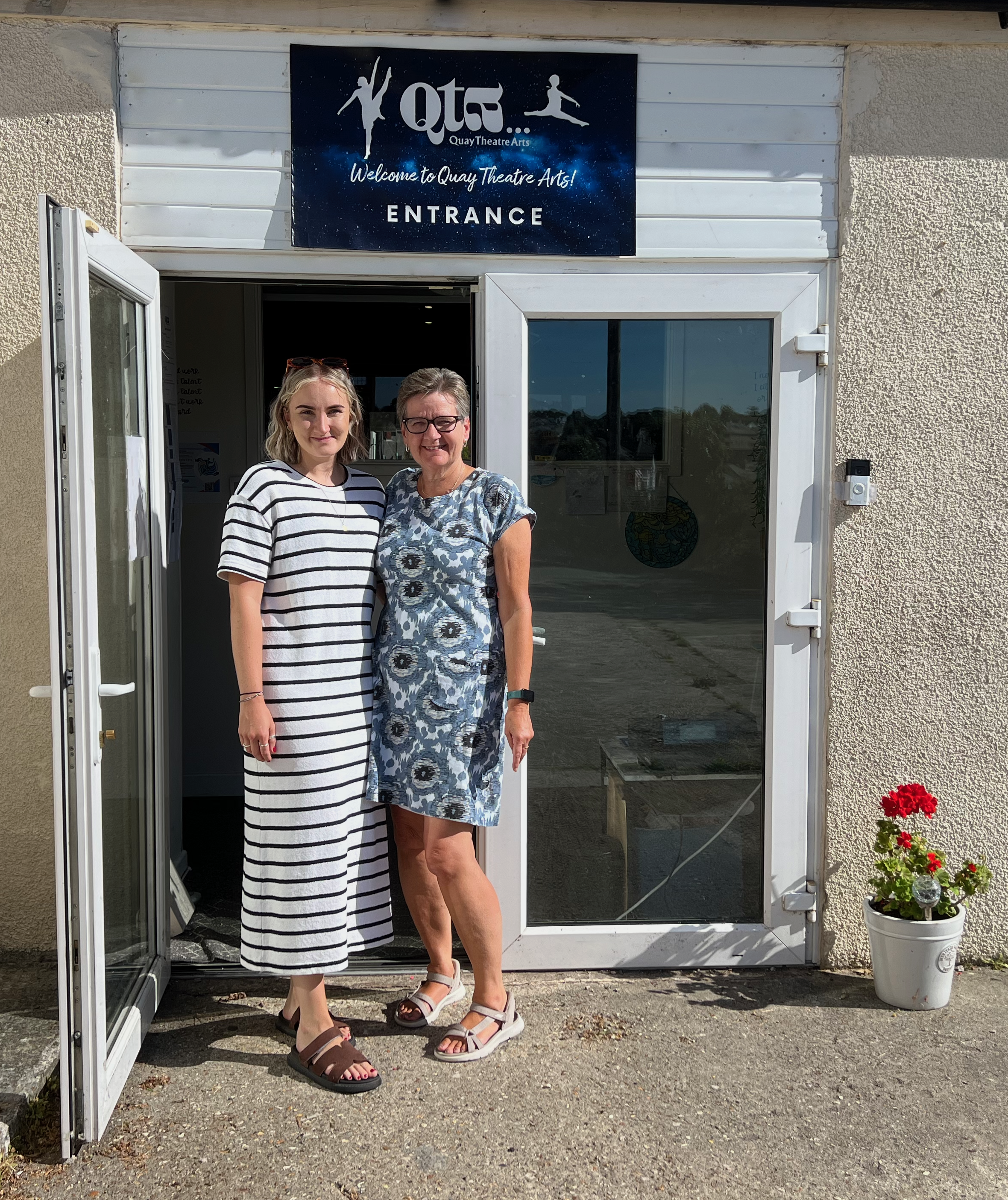 Two women standing in front of a glass door with a sign above that reads "Quay Theatre Arts." The woman on the left has shoulder-length blonde hair, wearing a black and white striped dress and sandals. The woman on the right has short gray hair, glasses, and is wearing a blue floral dress with sandals. The door has an open white frame, and there is a white flowerpot with red flowers on the right side of the door. The wall of the building is beige with a textured finish.