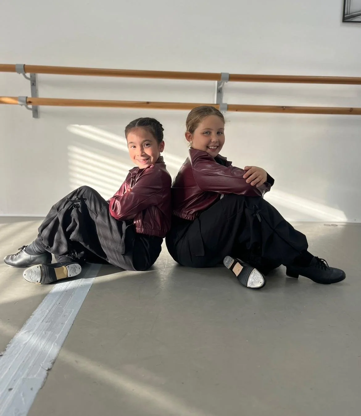 Two young girls in dance costumes sitting back-to-back on the floor of a dance studio, smiling at the camera, with ballet barres and a white wall in the background.
