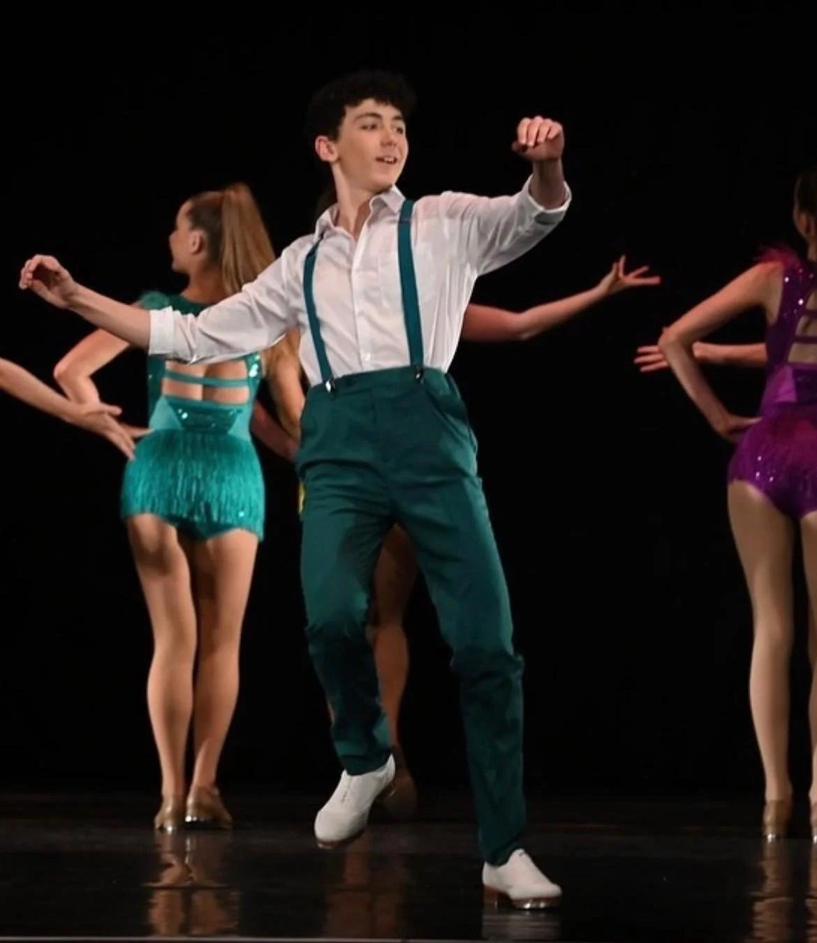 A young man performing a dance move on stage with several young women in colorful costumes dancing behind him.