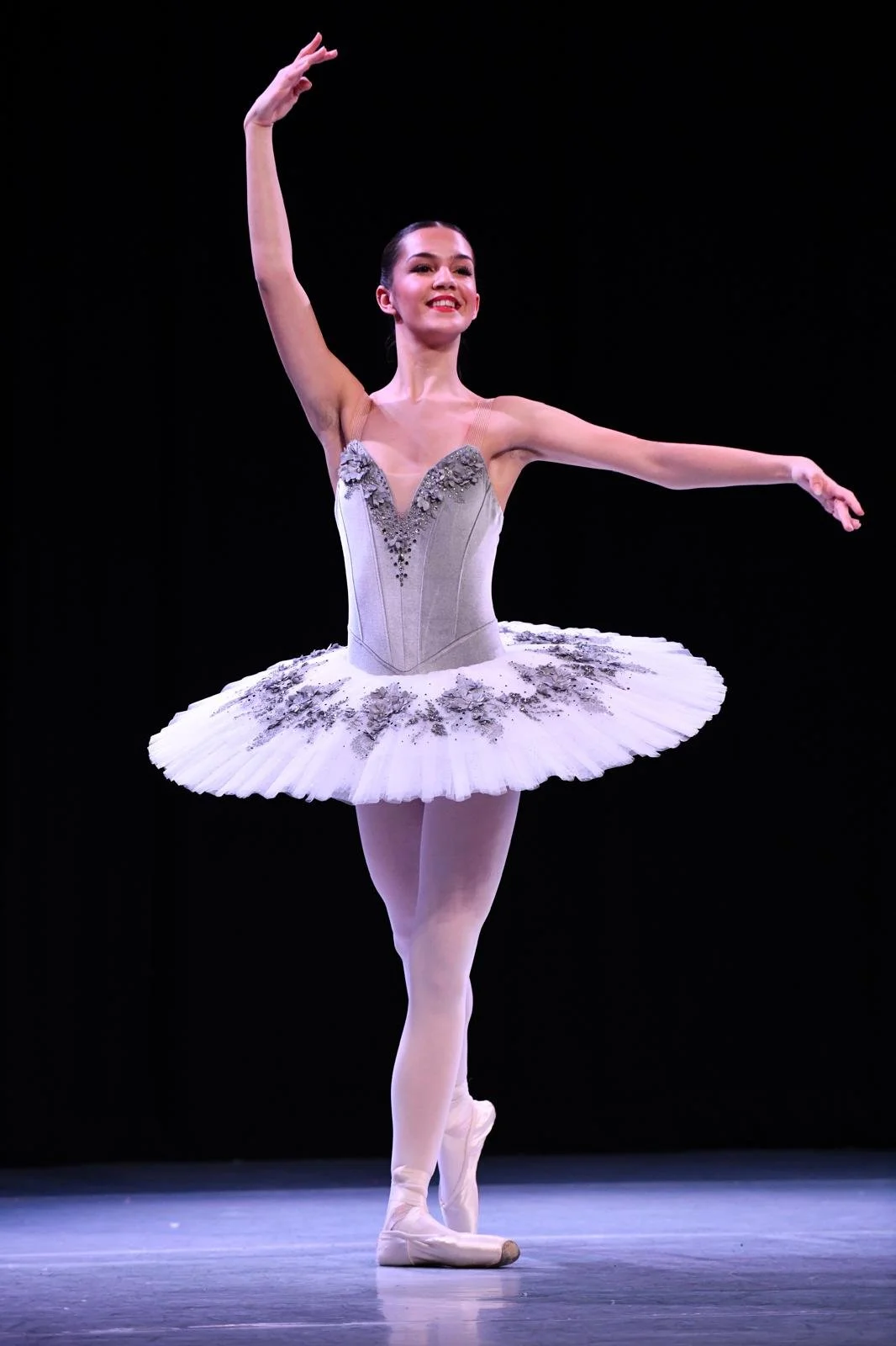 Ballet dancer wearing a white tutu with silver embellishments, performing on stage with a black background.