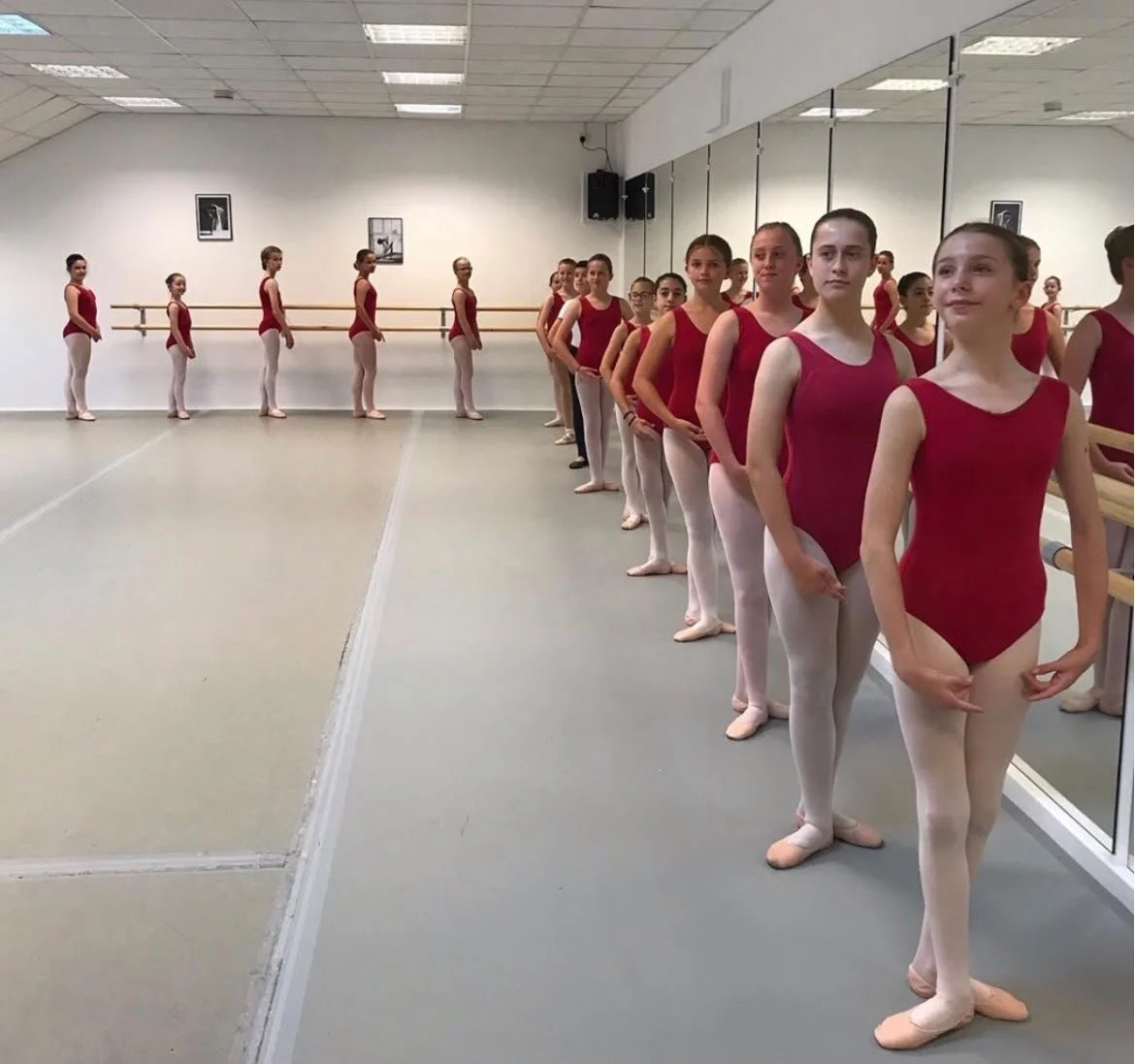 A group of young ballet dancers in red leotards and white tights standing in line in a dance studio, with some facing the mirror and others facing away, practicing ballet.