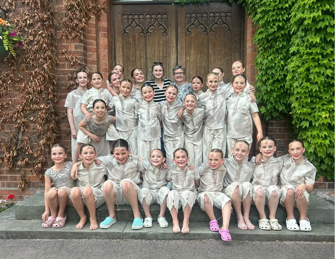 Group of young girls in matching light-colored outfits, posing with two adults in front of a wooden door surrounded by brick and greenery.
