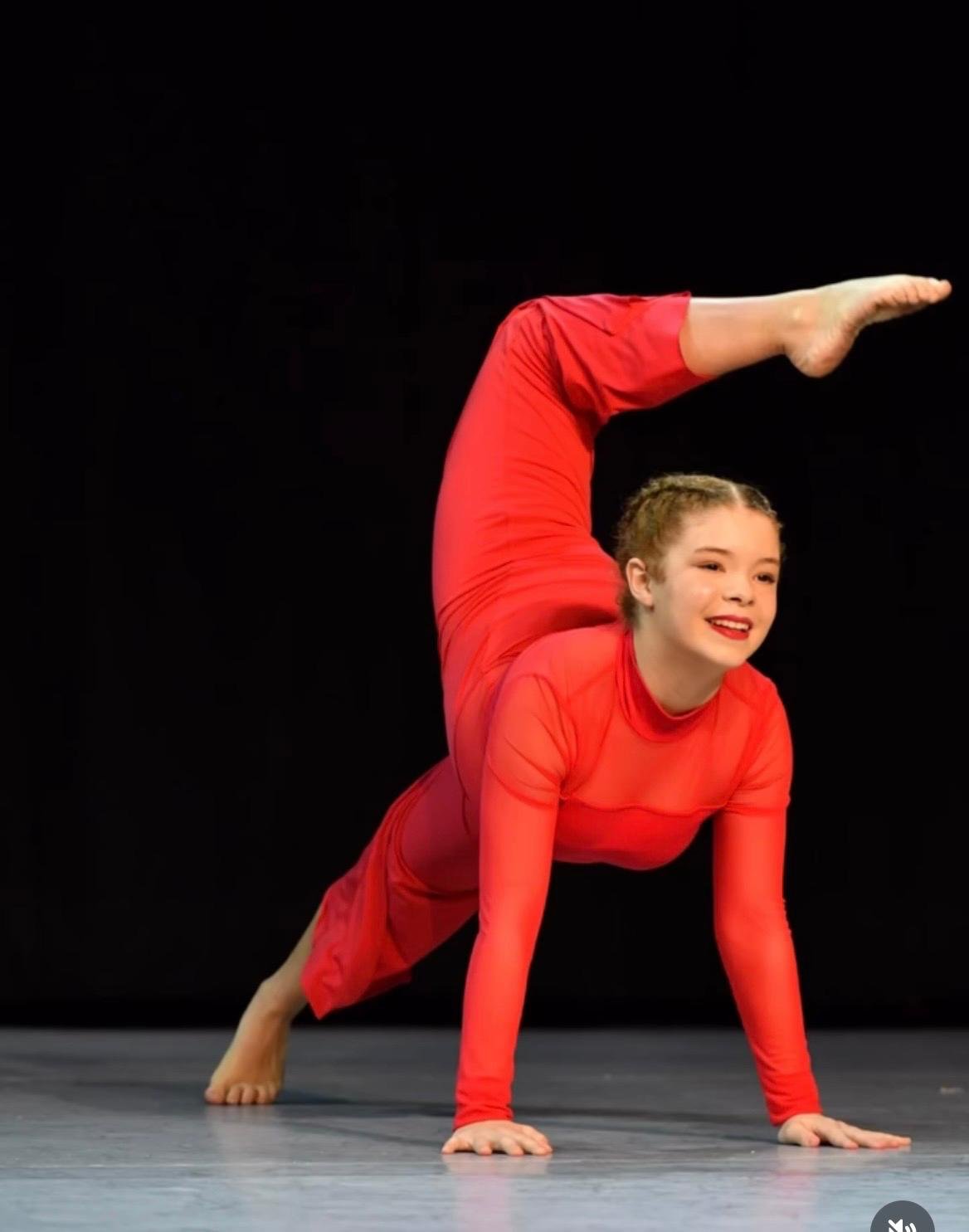 A young girl in a red dance costume balancing in a one-legged handstand position, with her other arm supporting her on the floor, against a black background.