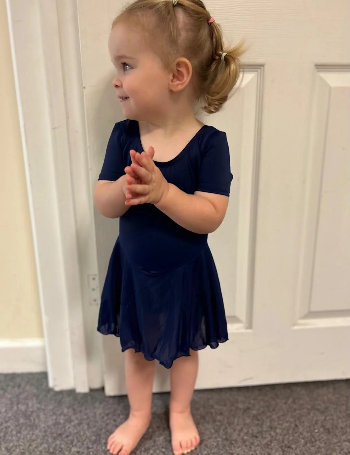A young girl with red hair tied in pigtails stands barefoot next to a closed white door, wearing a navy blue dress and smiling while looking to her left.
