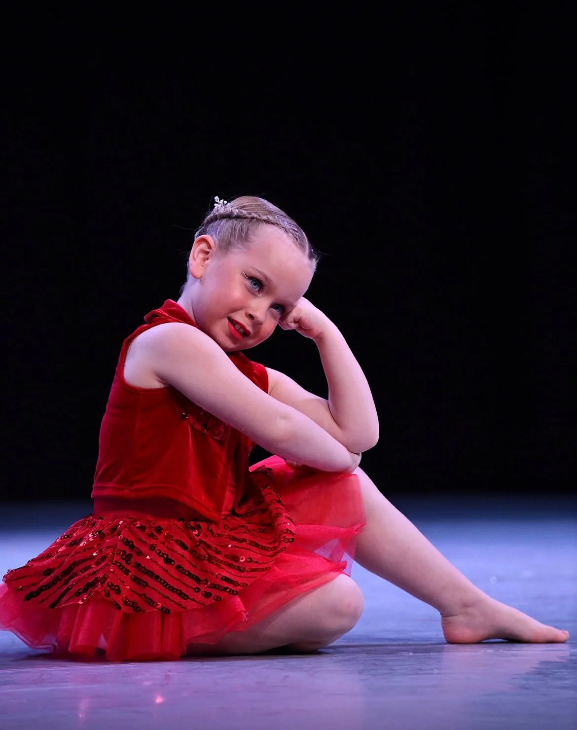 Young girl dressed in a red ballet costume, sitting on the floor with one leg extended and the other bent. She is resting her head on her hand and smiling at the camera, against a black background.