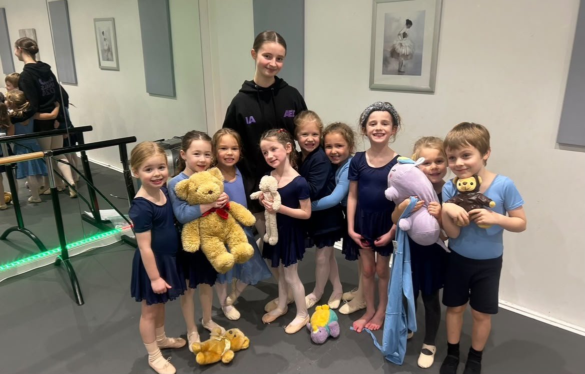 Group of young girls and a teenage girl posing with stuffed animals in a dance studio, with ballet shoes and hair buns, smiling at the camera.