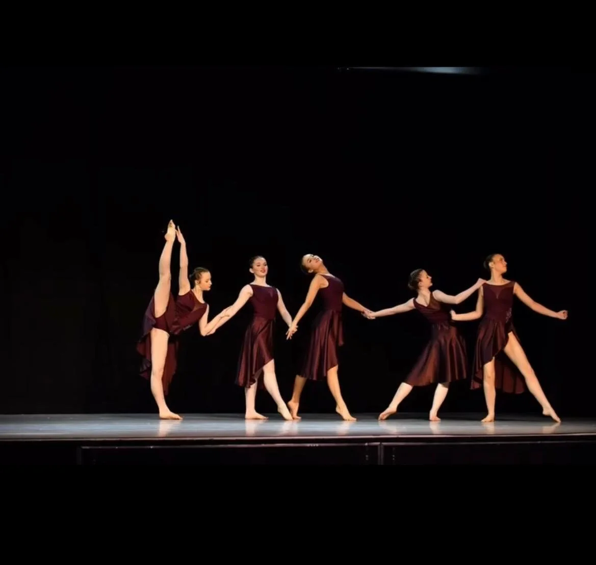 Six ballet dancers in burgundy costumes perform on stage against a black background, holding hands and striking poses.