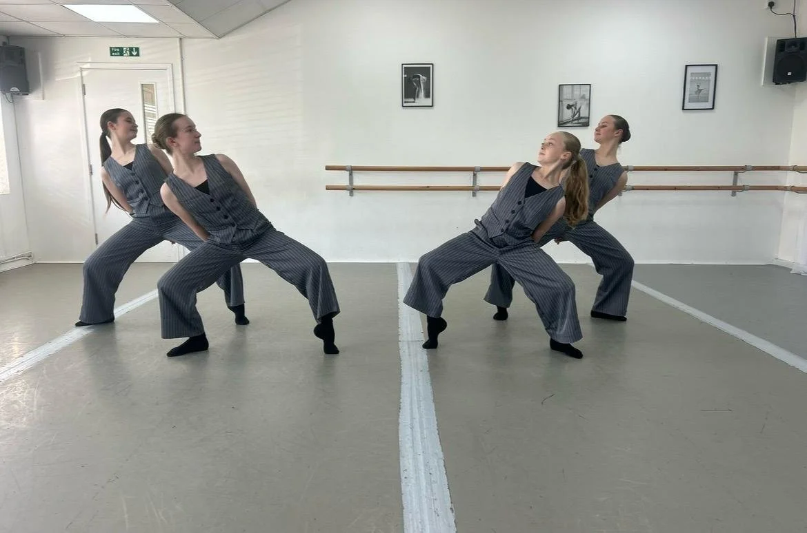 Four women in matching striped outfits practicing a dance or yoga pose in a dance studio with white walls, framed pictures, and a ballet bar.