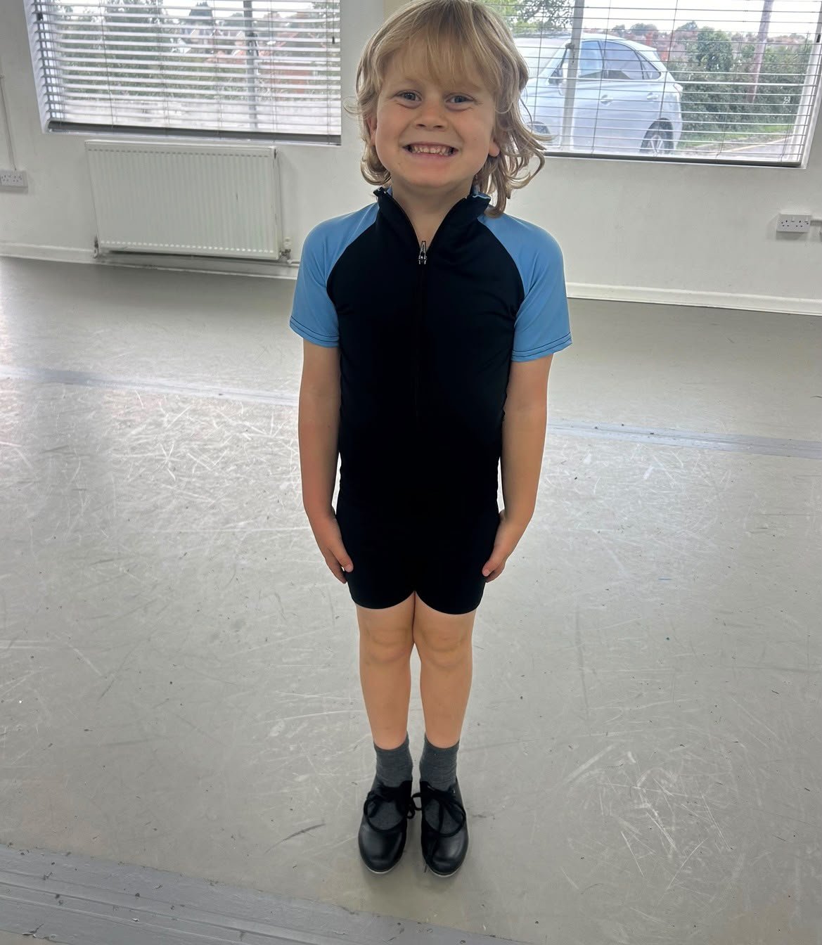 A young boy with blond hair smiling, standing indoors in a sports uniform, wearing black shoes, gray socks, and black shorts with a blue and black top.