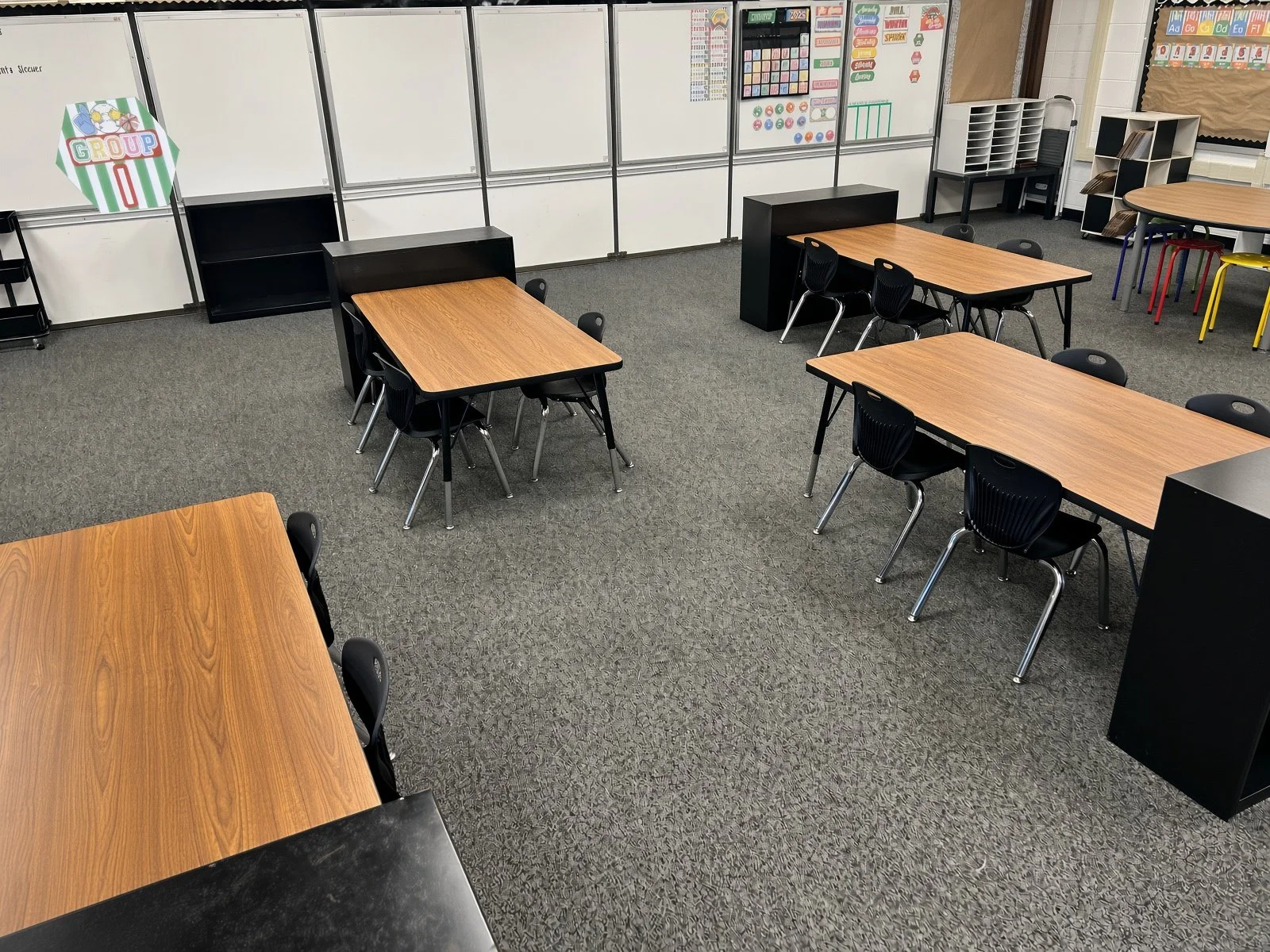 Elementary classroom with wooden tables, black chairs, and educational posters on a whiteboard, with a bulletin board in the background. Professionally installed and staged.
