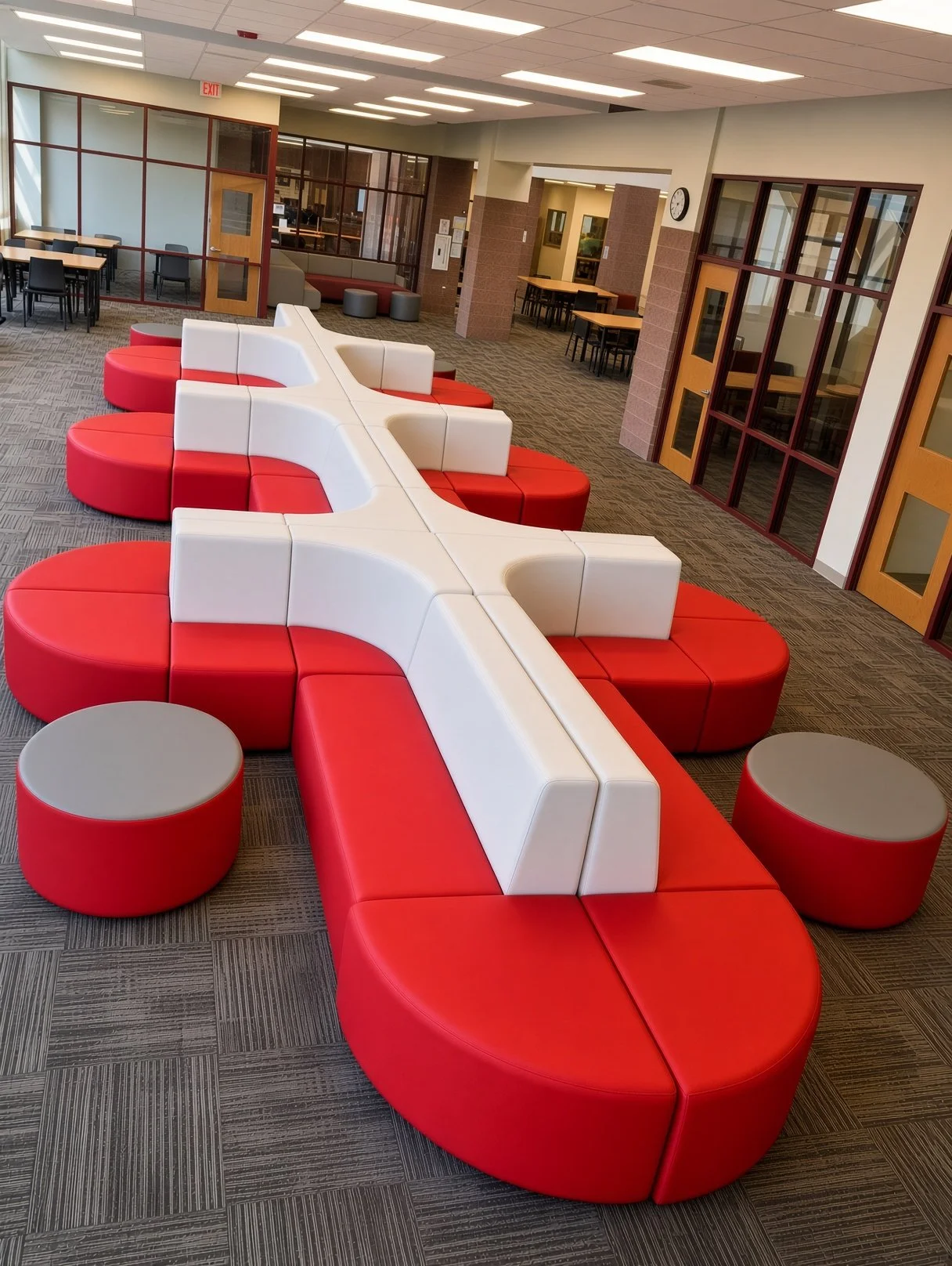 School library or common area with red and white modular seating arranged in a cross pattern, two round red ottomans, and a patterned gray carpet. There are multiple tables and chairs in the background.  Professionally staged and installed