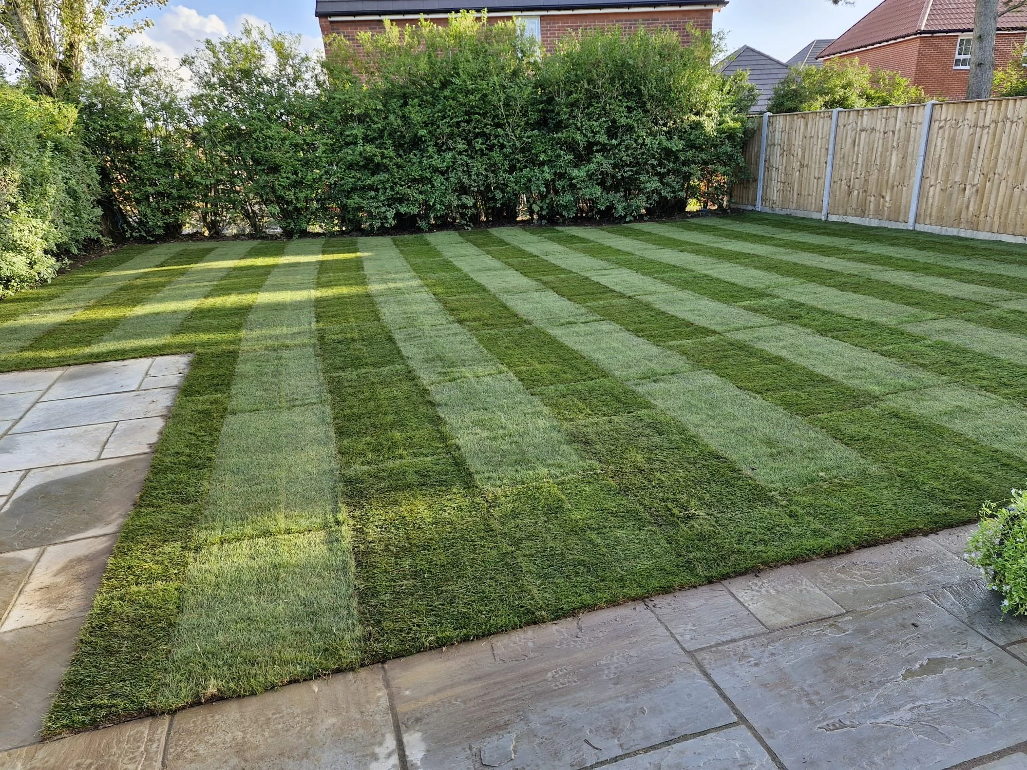 A backyard garden with freshly mowed grass showing light and dark green striped patterns, bordered by a brick house, a wooden fence, bushes, and trees.
