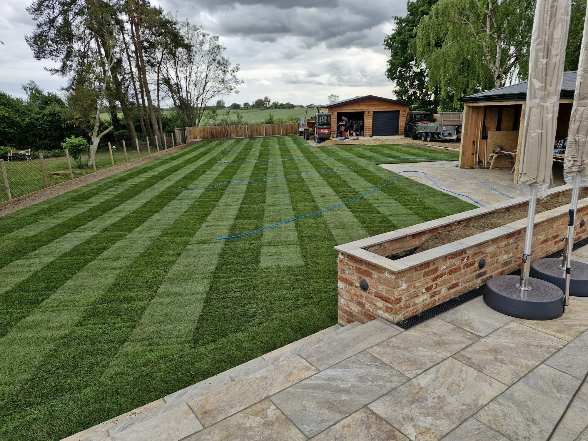 A backyard with freshly laid striped green grass, a wooden shed, outdoor furniture, and trees under a cloudy sky.