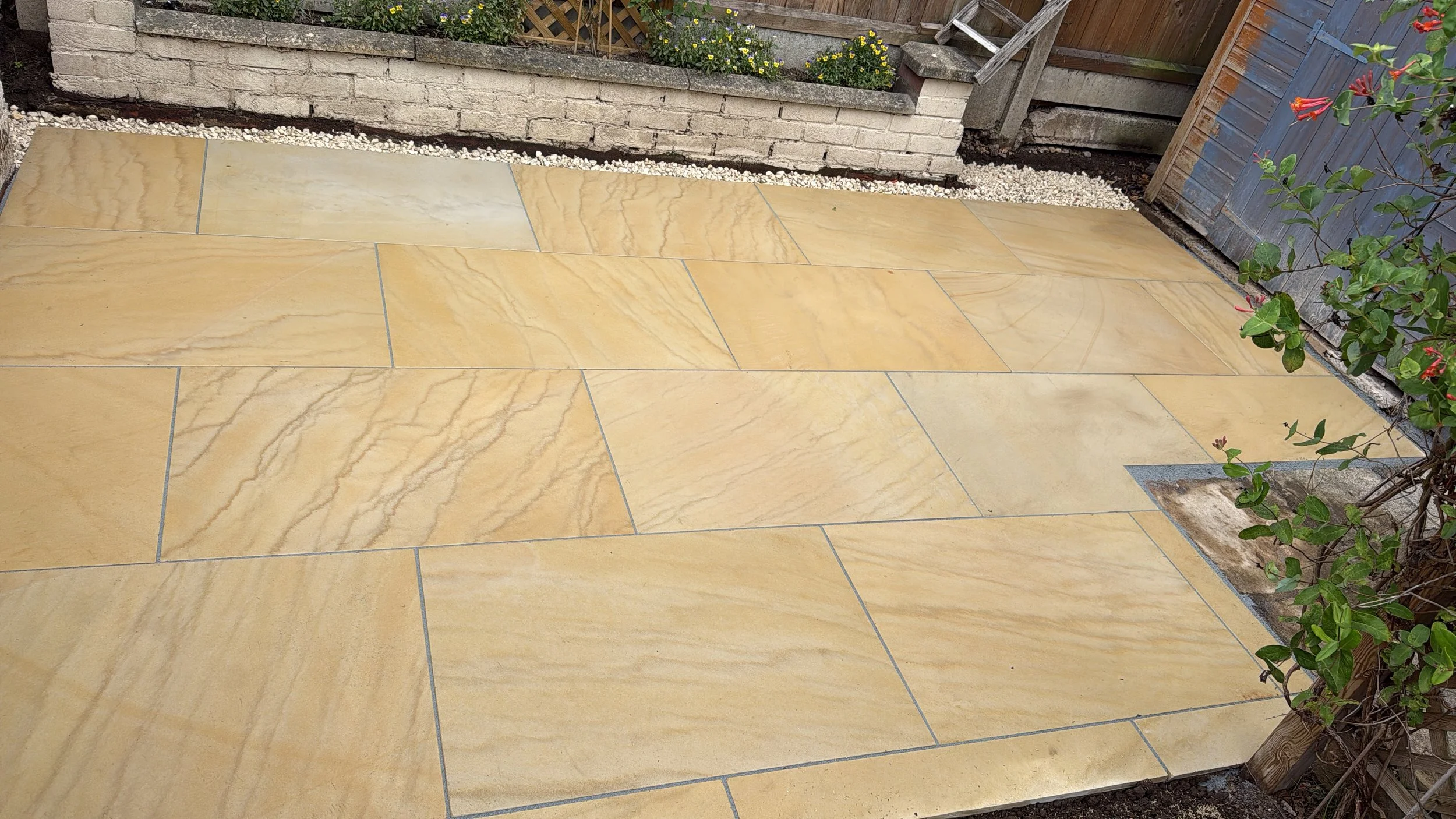 Yellow sandstone patio with garden flower bed in the background, bordered by decorative white gravel, and a wooden fence with climbing plants on the right side.