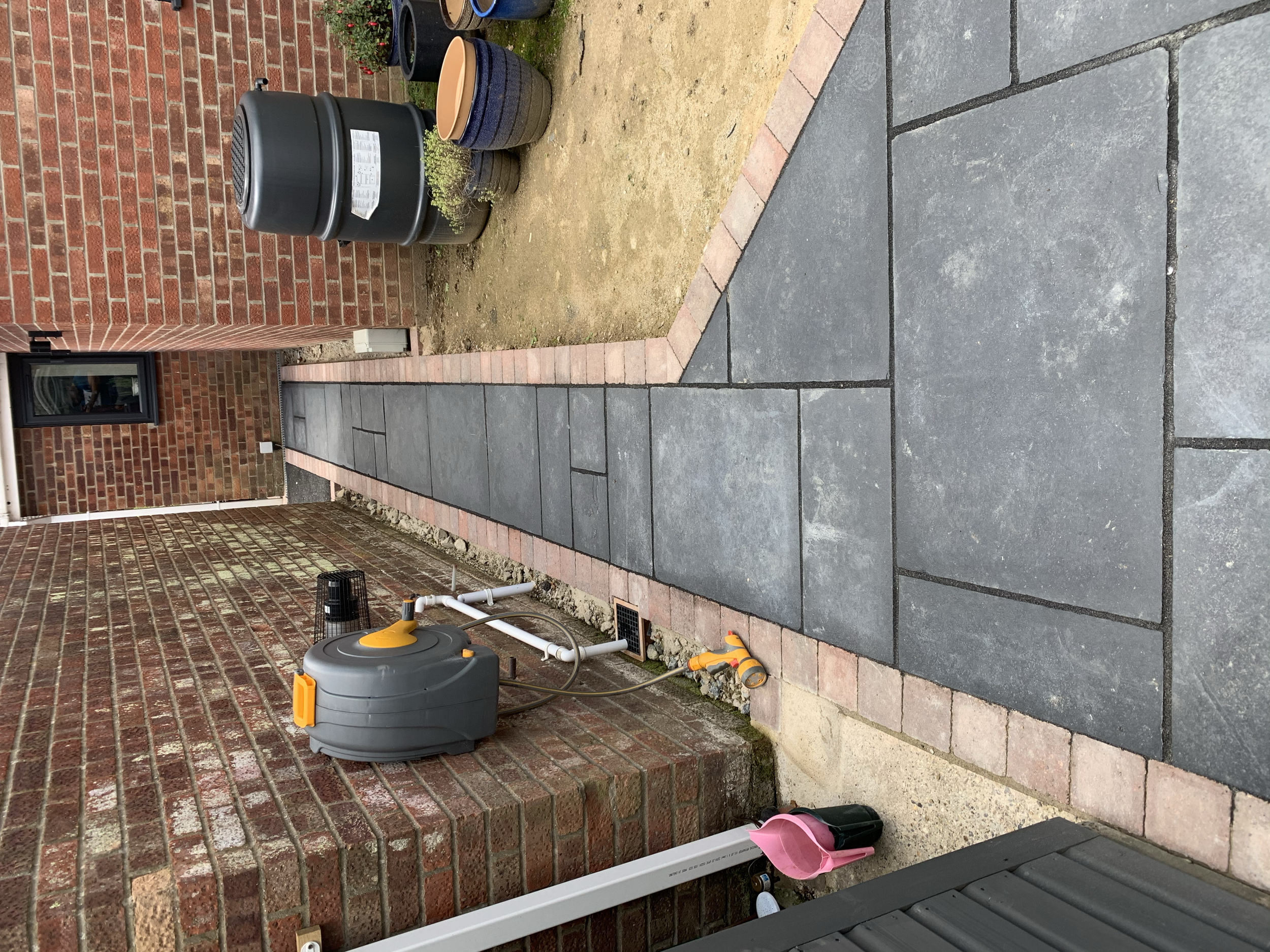 A newly paved stone pathway runs between brick walls of a house exterior, with plants and flower pots on the right side, and a wall-mounted hose reel and garden tools on the left.