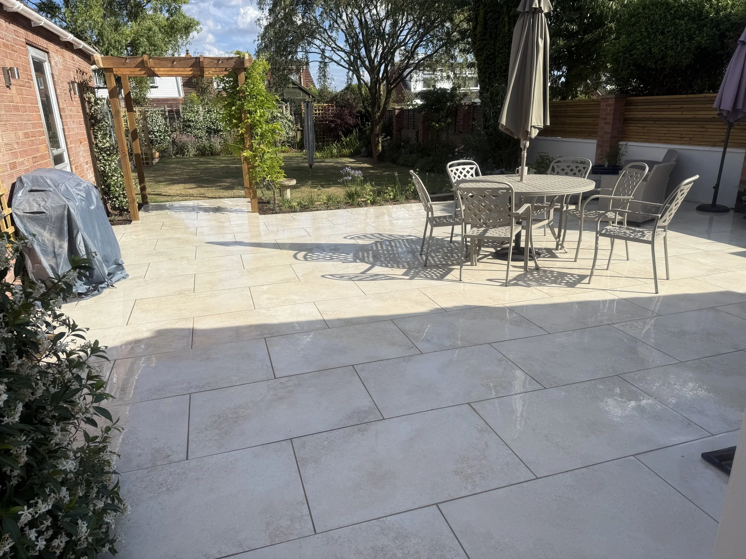 A backyard patio with white tile flooring, outdoor furniture, a patio umbrella, and a garden with trees and bushes.