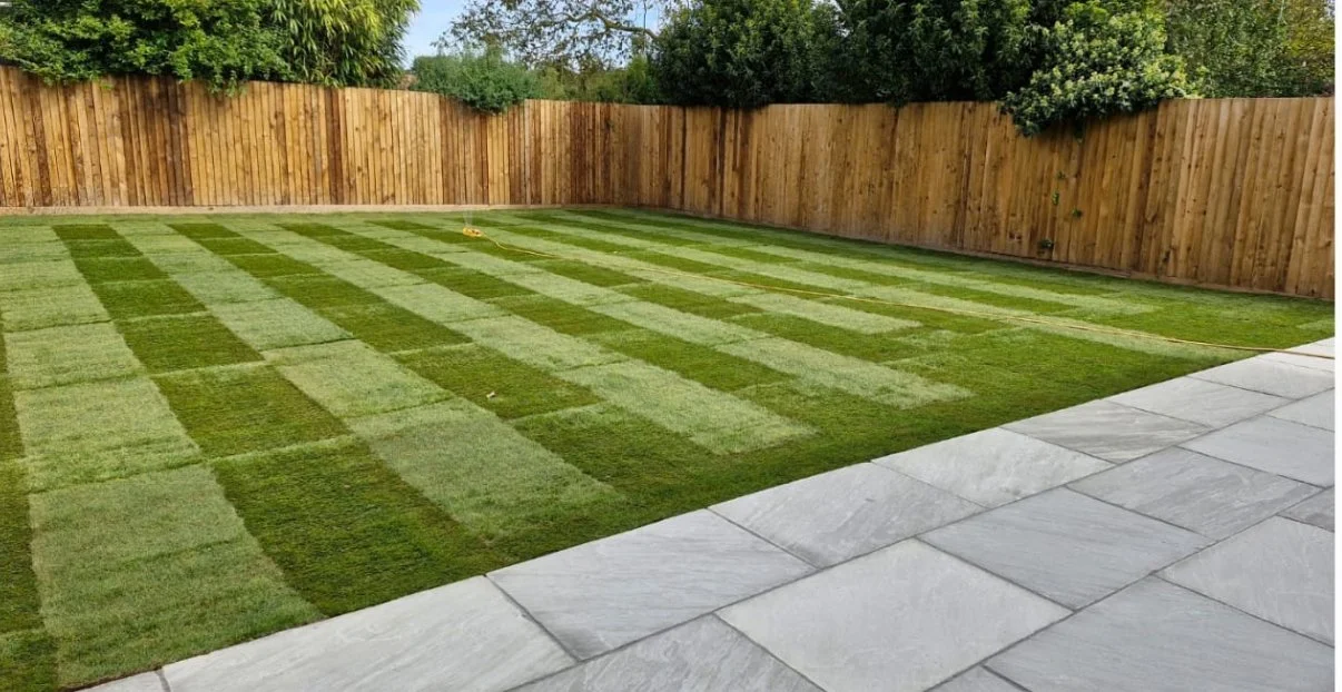 A backyard with newly laid sod grass arranged in a checkerboard pattern, surrounded by a wooden privacy fence and a concrete patio in the foreground.