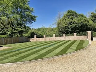 Well-maintained lawn with green and yellow stripes, bordered by a gravel pathway and a brick wall with pillars, under a blue sky with trees in the background.