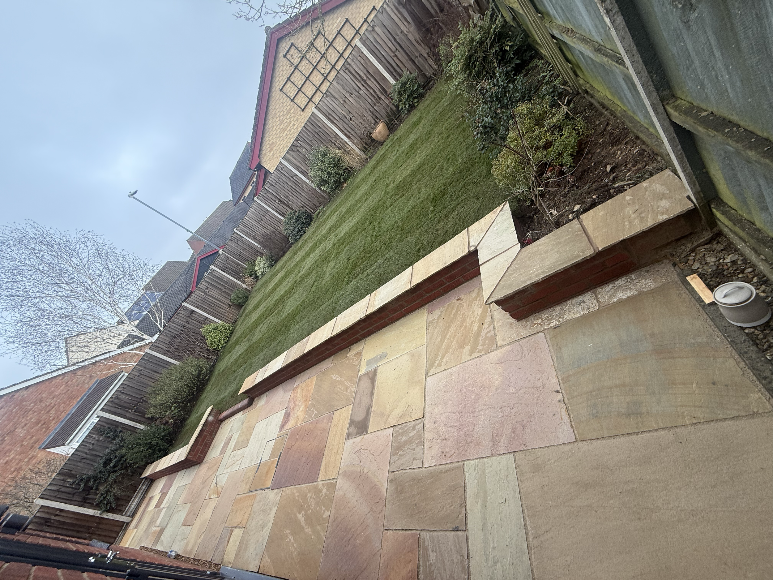 A backyard with a patio made of multicolored stone tiles, a grassy lawn with small bushes, a wooden fence, and a brick shed with a metal trellis on the side. There is a nearby streetlamp and an overcast sky.