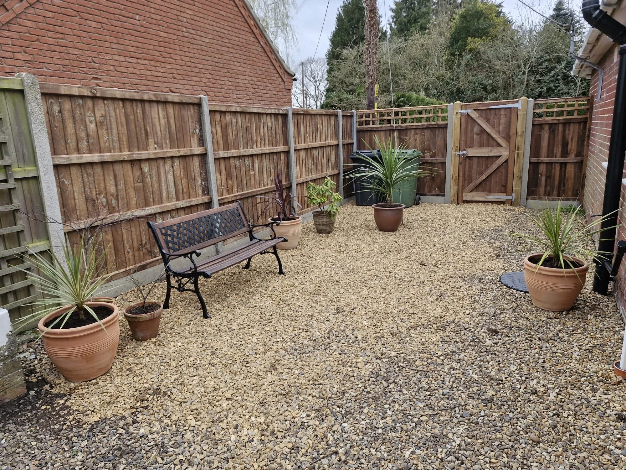 A backyard with a gravel ground, potted plants, a wooden and concrete fence, a garden bench, and some trees in the background.