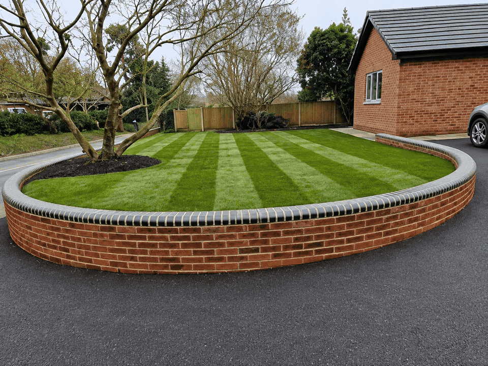 A brick garden bed with a curved shape, containing a tree and lawn with alternating light and dark green stripes, adjacent to a house with a brick exterior and a car parked nearby.